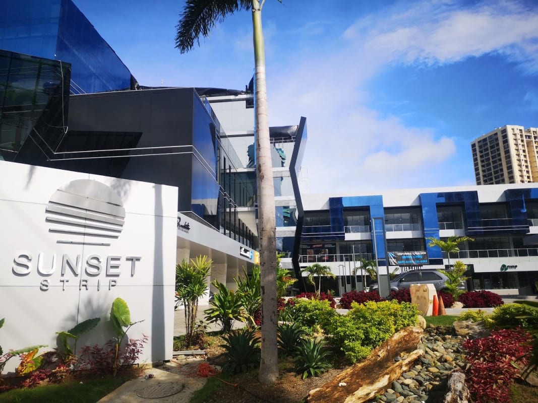 Angular glass commercial center Sunset Strip with garden, palm trees, and signage Via Israel Panama