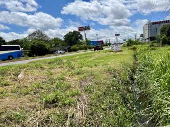 Open vacant plot of land with gravel, grass patches, puddle, trees, and blue sky in commercial area David Chiriquí Panama