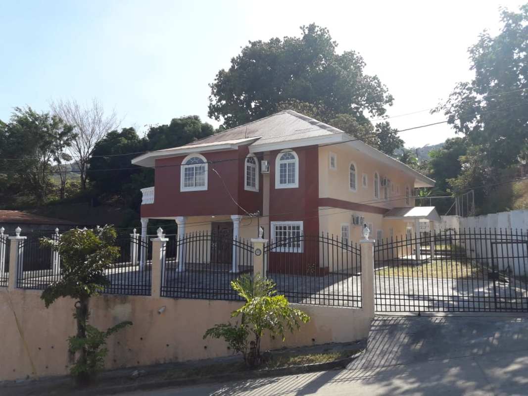 Two-story cream colored house with large paved driveway gated fence and tropical garden in Las Cumbres Panama for sale