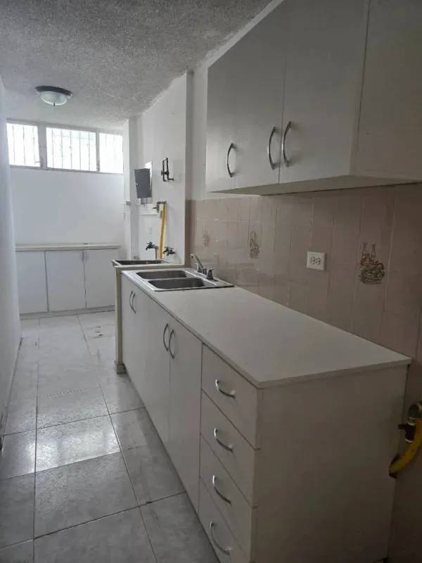 Narrow minimalist kitchen with dual sink, white cabinets, beige tiles and barred window in PH La Alameda Panama