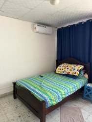 Simple tiled bedroom with wooden bed, colorful bedding, blue curtains, AC mounted in Chitré residence.
