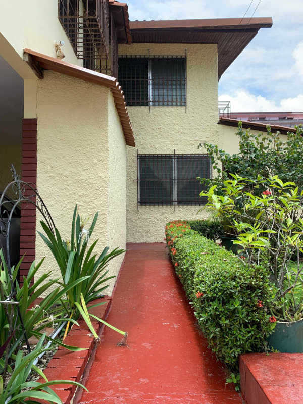 Garden walkway with shrubs leading to a cream house with red accents and security bars in La Loceria Panama